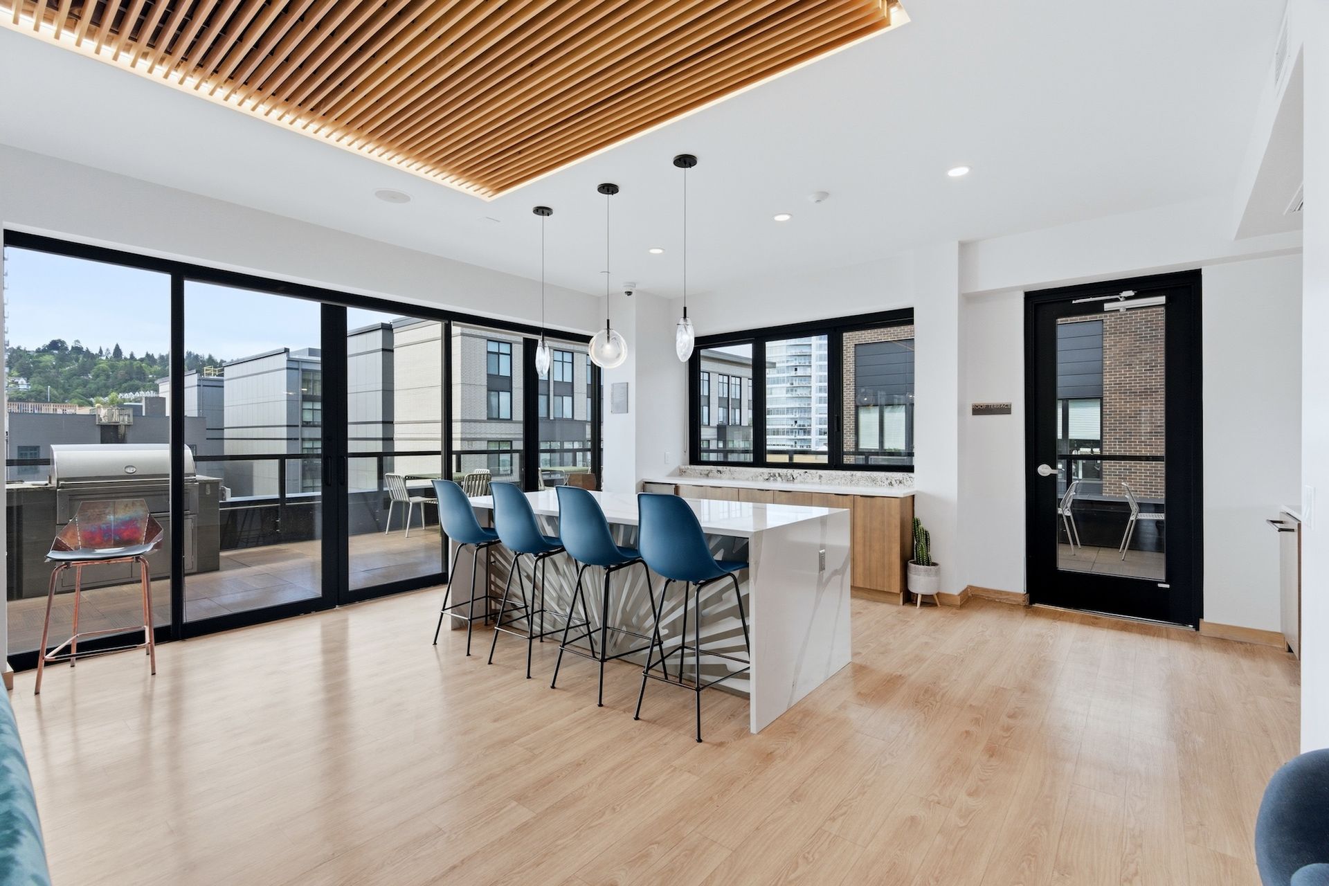 Modern open-plan kitchen with island, blue bar stools, light wood floors, and large windows overlooking a patio.