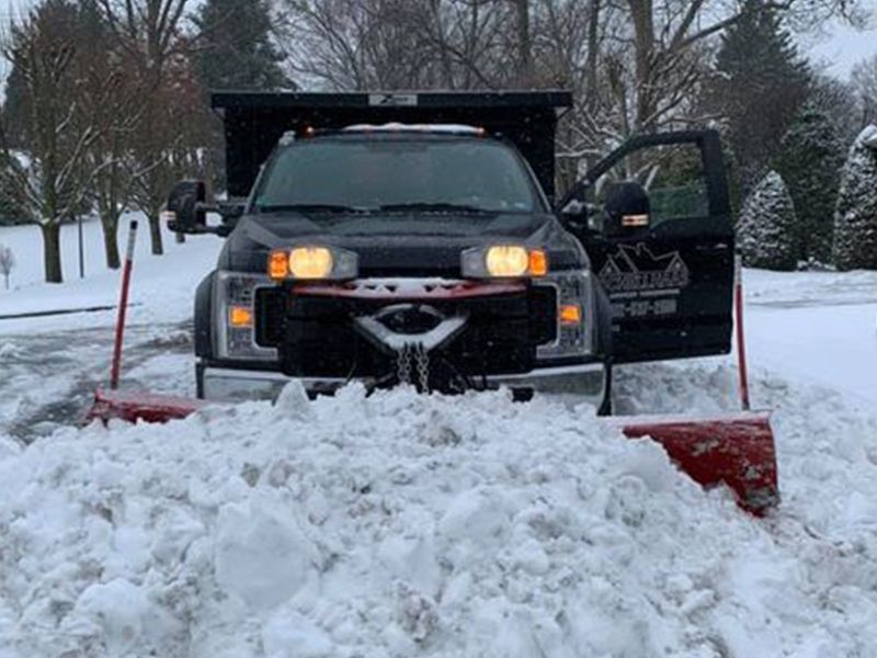 Black snowplow clearing deep snow on a residential street, headlights on, driver door open.
