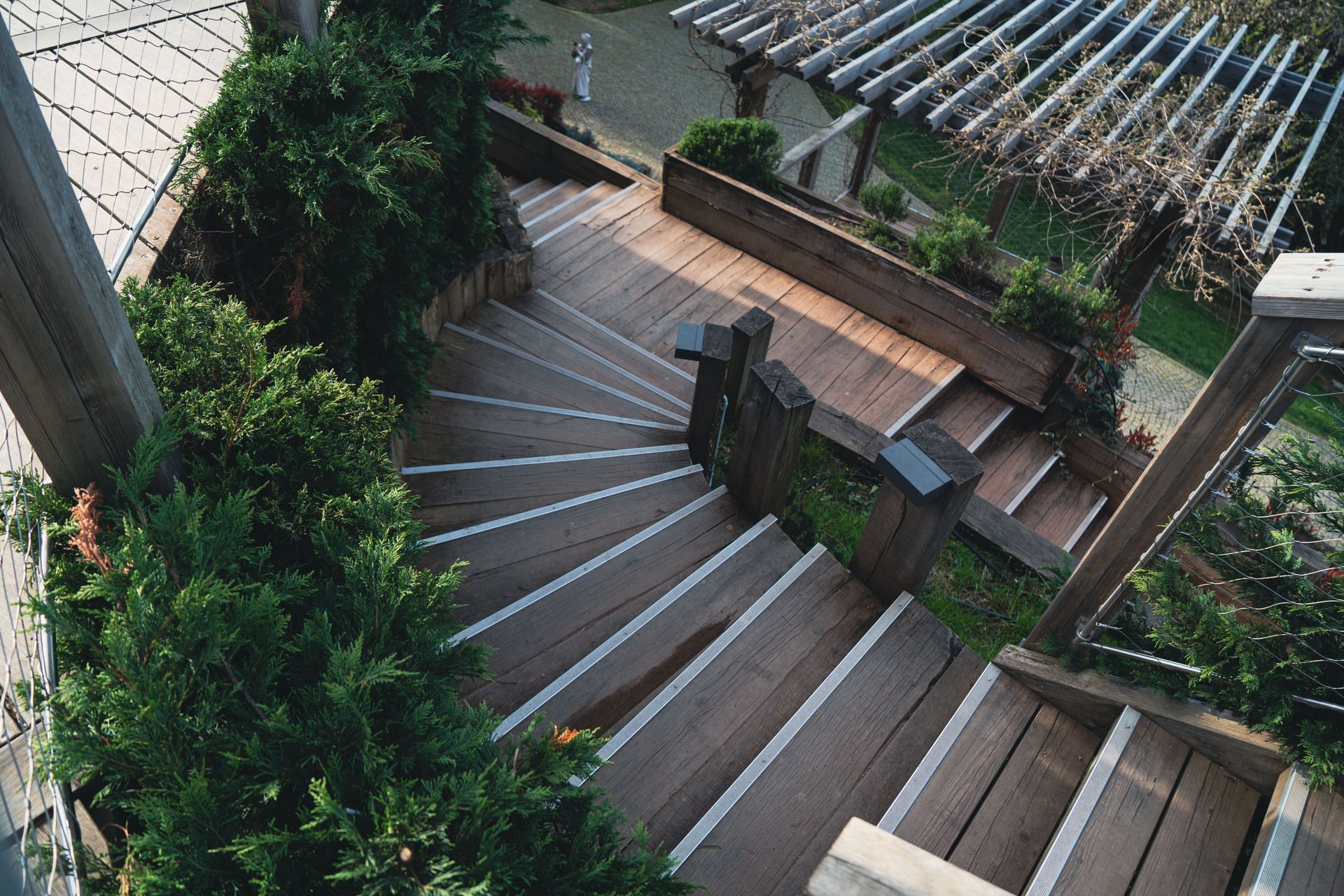Curved wooden staircase beside lush greenery and a terraced walkway, viewed from above.