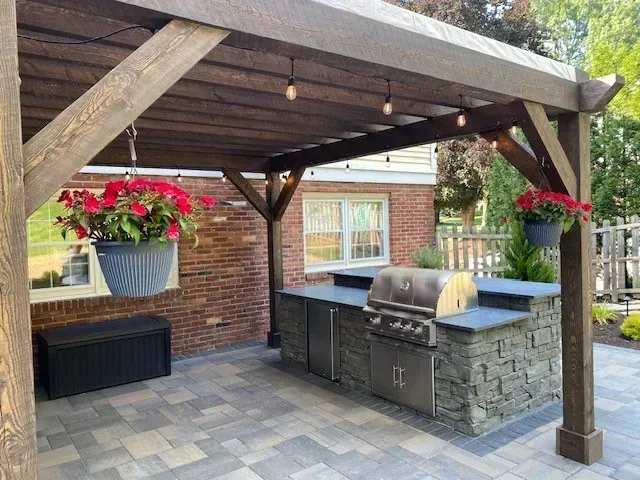 Backyard patio with wooden pergola, stone outdoor kitchen, grill, and hanging red flowers