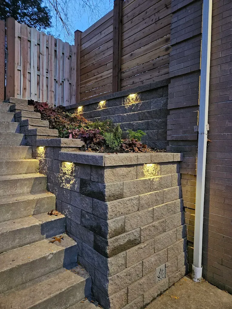 Outdoor stone stairs with wall lights, gray retaining wall, and wooden fencing at dusk