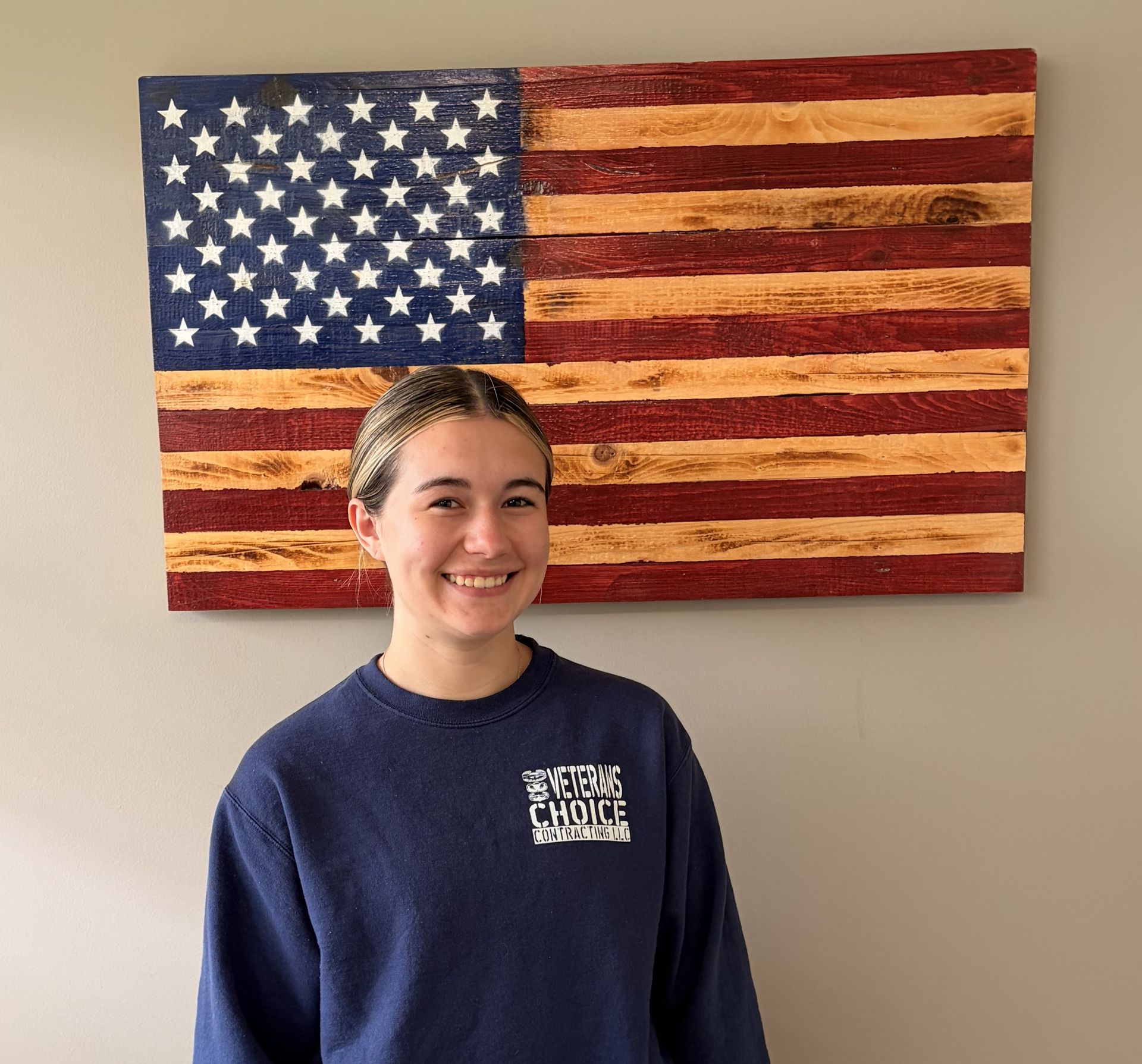 Smiling person in a navy shirt standing before a wooden American flag wall hanging