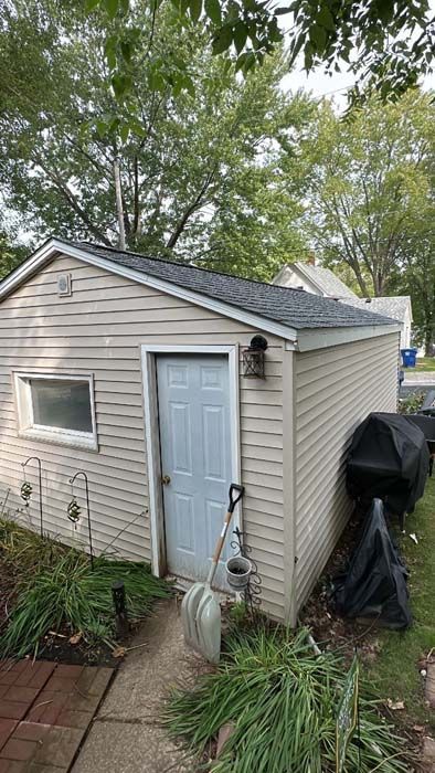 Tan shed with gray roof, light blue door, and small window; shovel leans against the door.