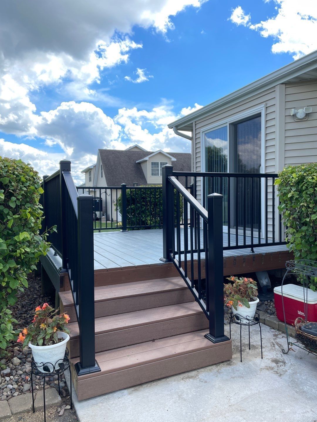 Deck with black railing, steps, and composite decking, next to a house under a cloudy blue sky.