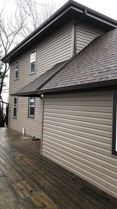 Two-story house with tan siding, brown roof and gutters. Weathered wooden deck in the foreground.