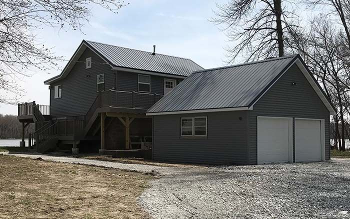 Gray house with attached garage, dark metal roofs, and wooden deck. Gravel driveway.