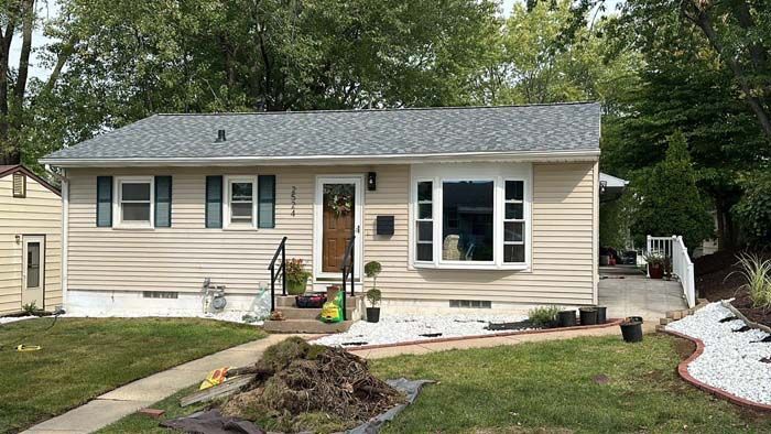 Tan house with grey roof and shutters. Lawn in front with some landscaping, walkway and shrubs.