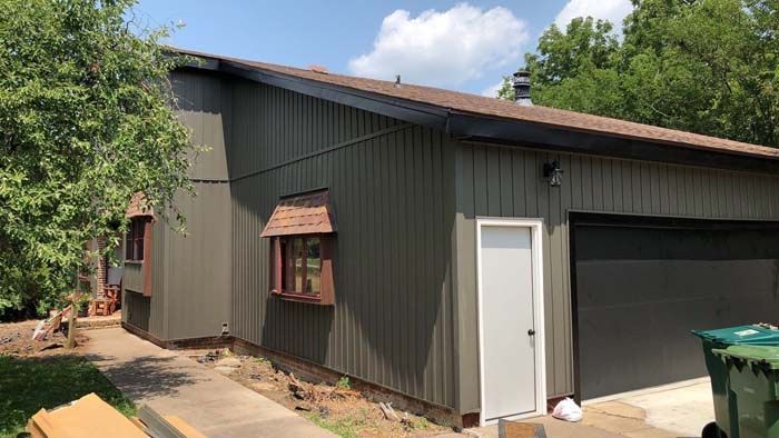 Exterior of a house with dark green siding, brown trim, and a garage door. A small white door is visible.