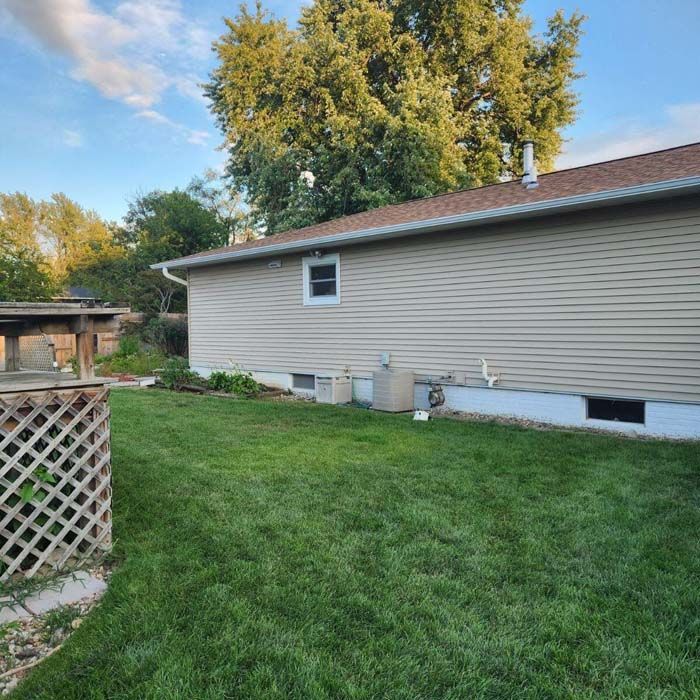 Backyard with a beige house, green lawn, lattice fence, and trees under a partly cloudy sky.