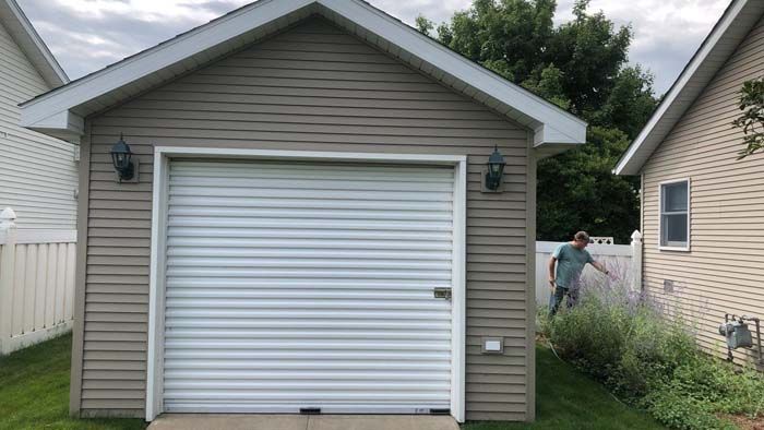 Tan garage with white roll-up door, man trimming tall grass next to it.