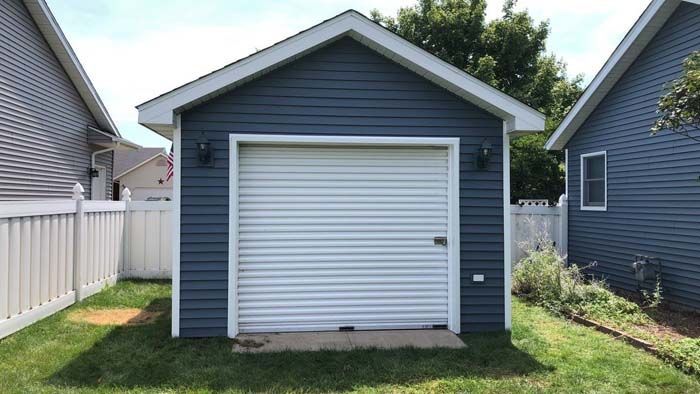 Blue garage with white roll-up door, lights, and siding. Located between two houses with white fences.