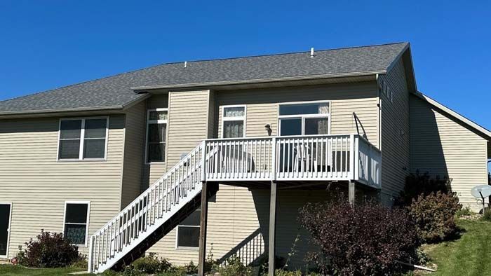 Back of a two-story beige house with a gray shingled roof, deck, and white stairs against a blue sky.