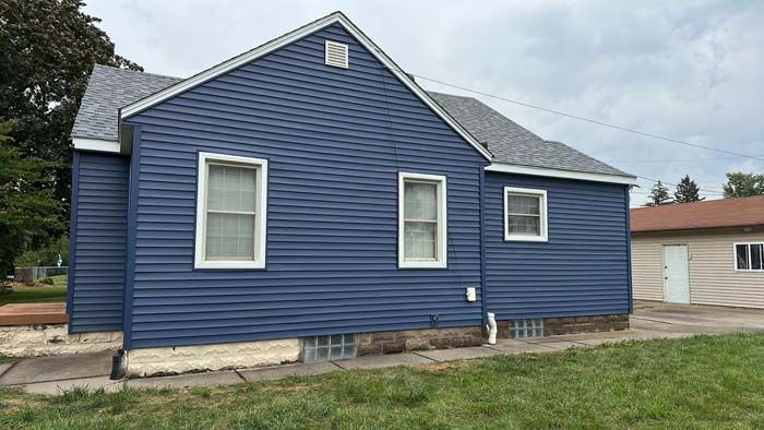 Blue-sided house with three white-framed windows, gray roof, and a beige garage in the background.