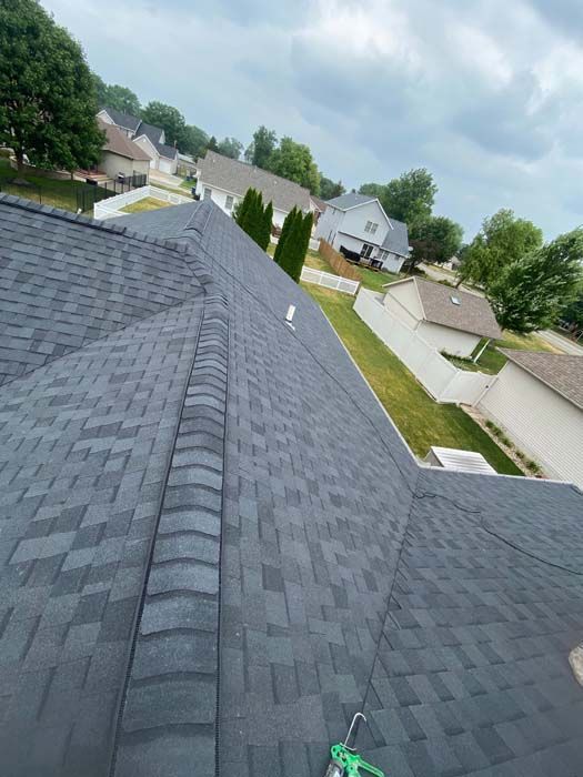 Dark grey shingled roof with a tool resting on it. Houses and trees are in the background, under an overcast sky.