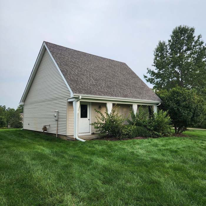 Small beige building with a gabled roof and a porch. Surrounded by green grass and a cloudy sky.