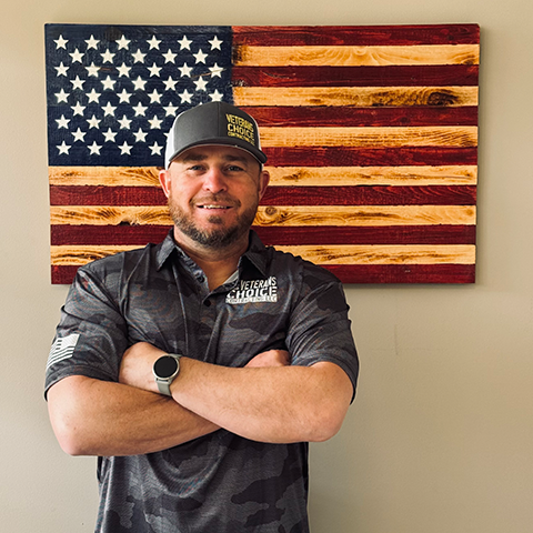 Man with arms crossed smiling in front of a wooden American flag. He is wearing a cap and camo shirt.