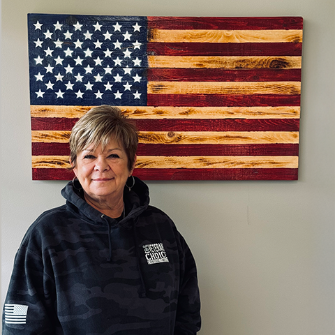 Woman standing in front of wooden American flag. She wears a black hooded sweatshirt.
