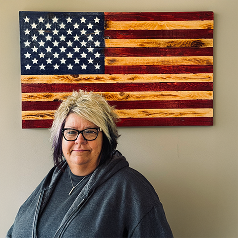 Woman with glasses, gray hoodie, stands in front of a wooden American flag.
