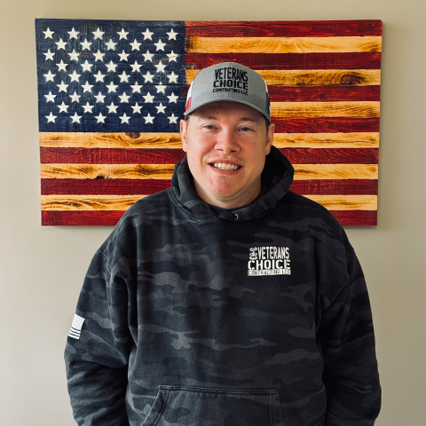 Man in camouflage hoodie and cap smiles in front of a wooden American flag.
