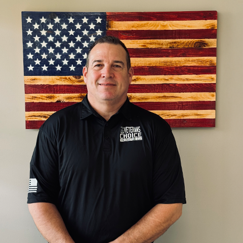 Man in black polo shirt stands in front of a wooden American flag.