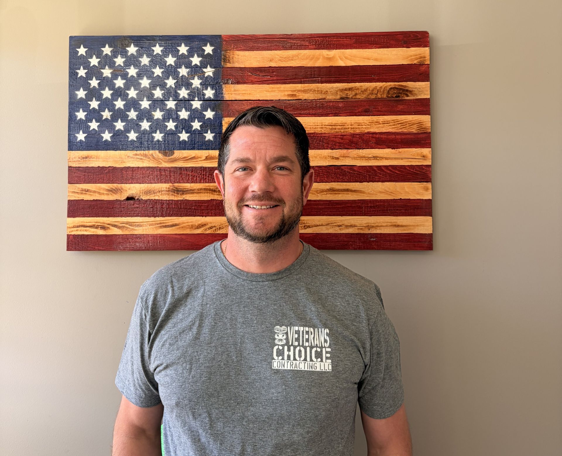 Man standing indoors before a painted American flag, wearing a gray T-shirt and smiling.