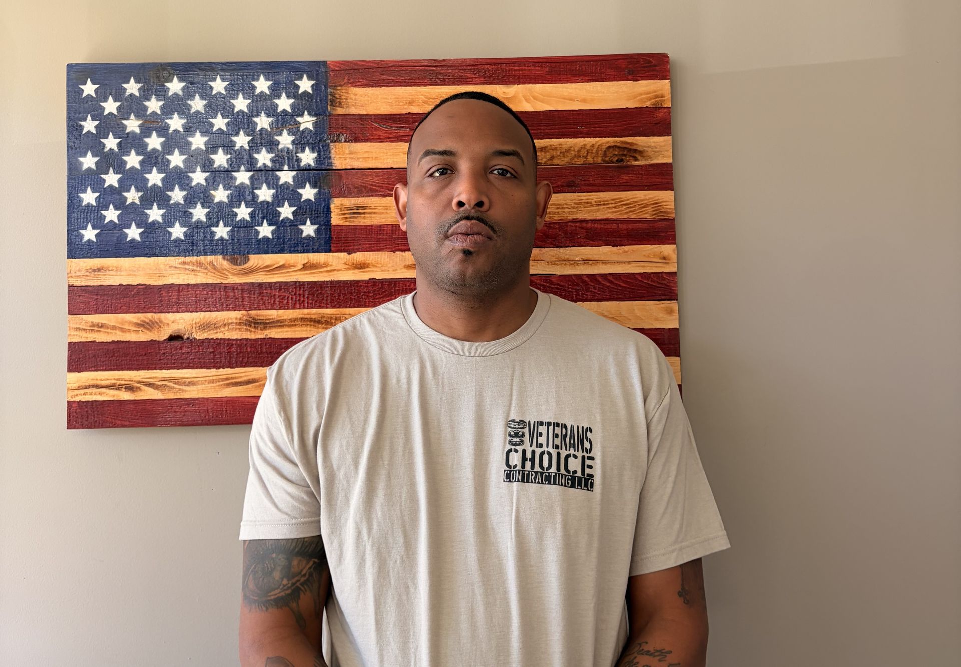 Man standing indoors in front of a wooden American flag wall art, wearing a beige T-shirt.