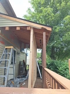 Covered porch under construction with ladder, tools, and wooden railing beside trees