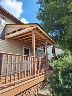 Wooden porch with railing and sloped roof on a beige house, framed by trees and blue sky