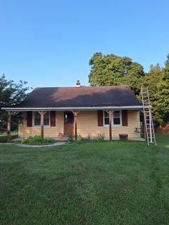 Single-story beige house with a dark roof, front porch, green lawn, and ladder by the right side.