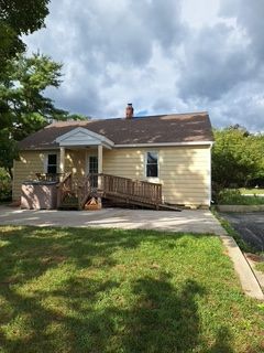 Small single-story house with a ramp, brick steps, driveway, and green front lawn under cloudy skies