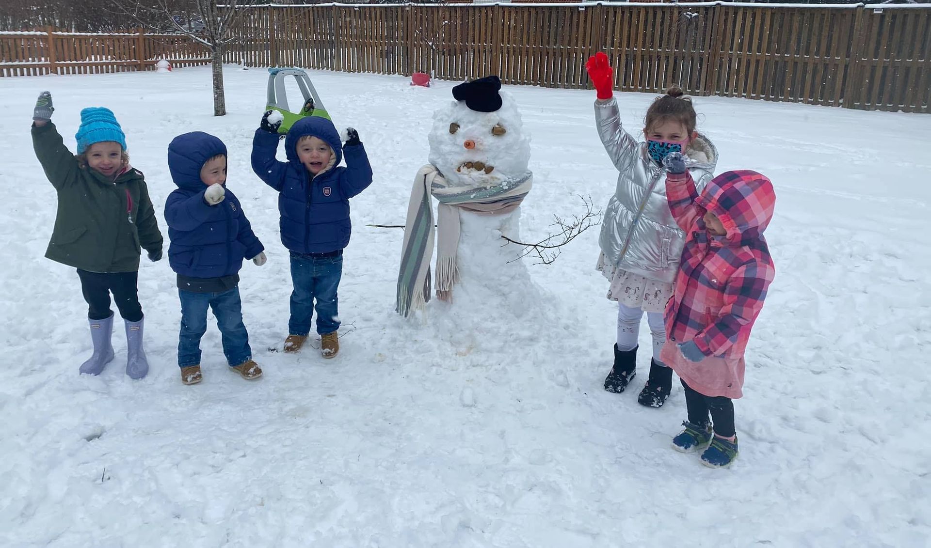 Children in winter clothes stand beside a snowman in a snowy park, waving and smiling.