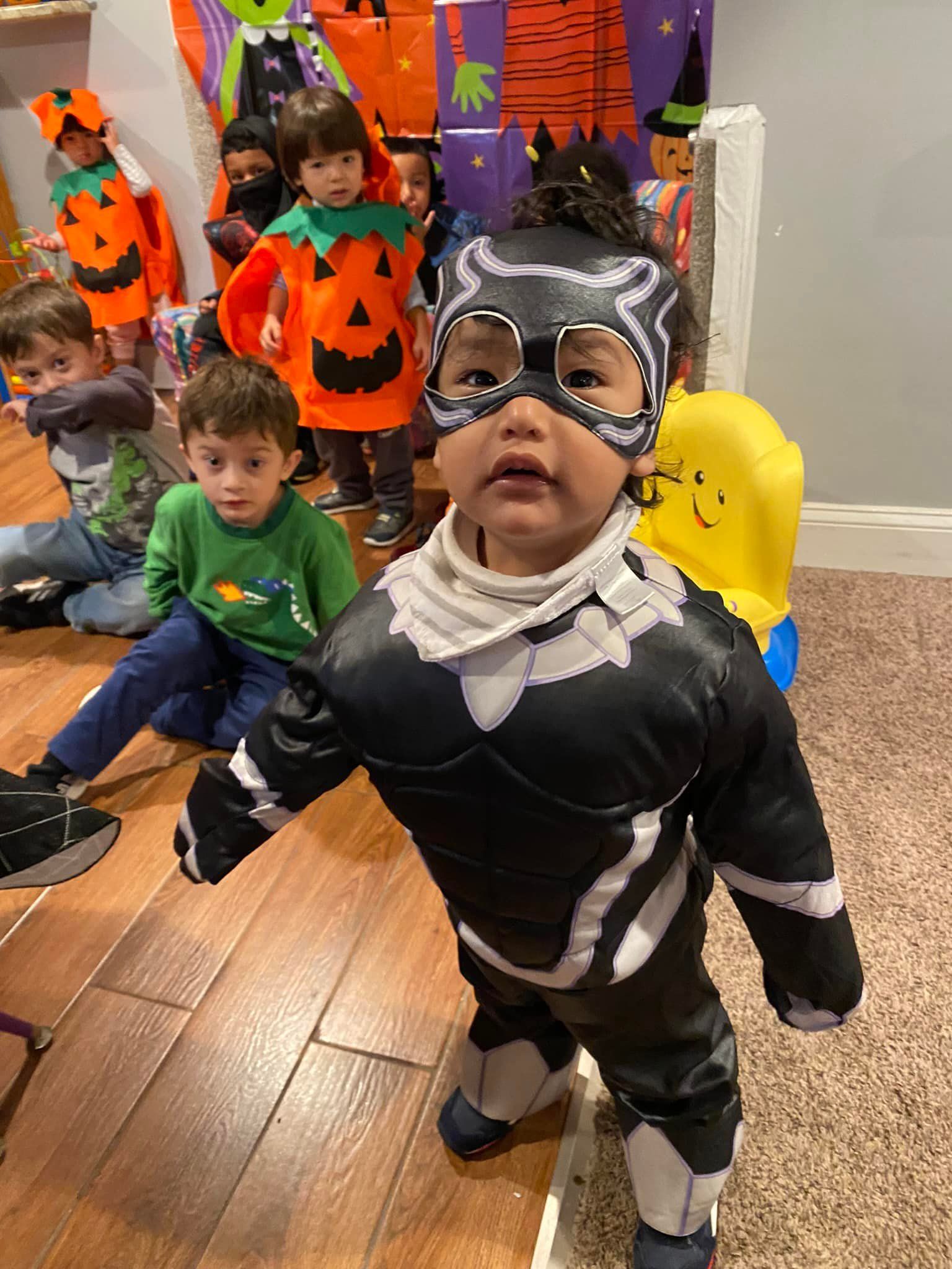 Toddler in black-and-white costume and silver mask at a Halloween party, with children in pumpkin outfits behind.