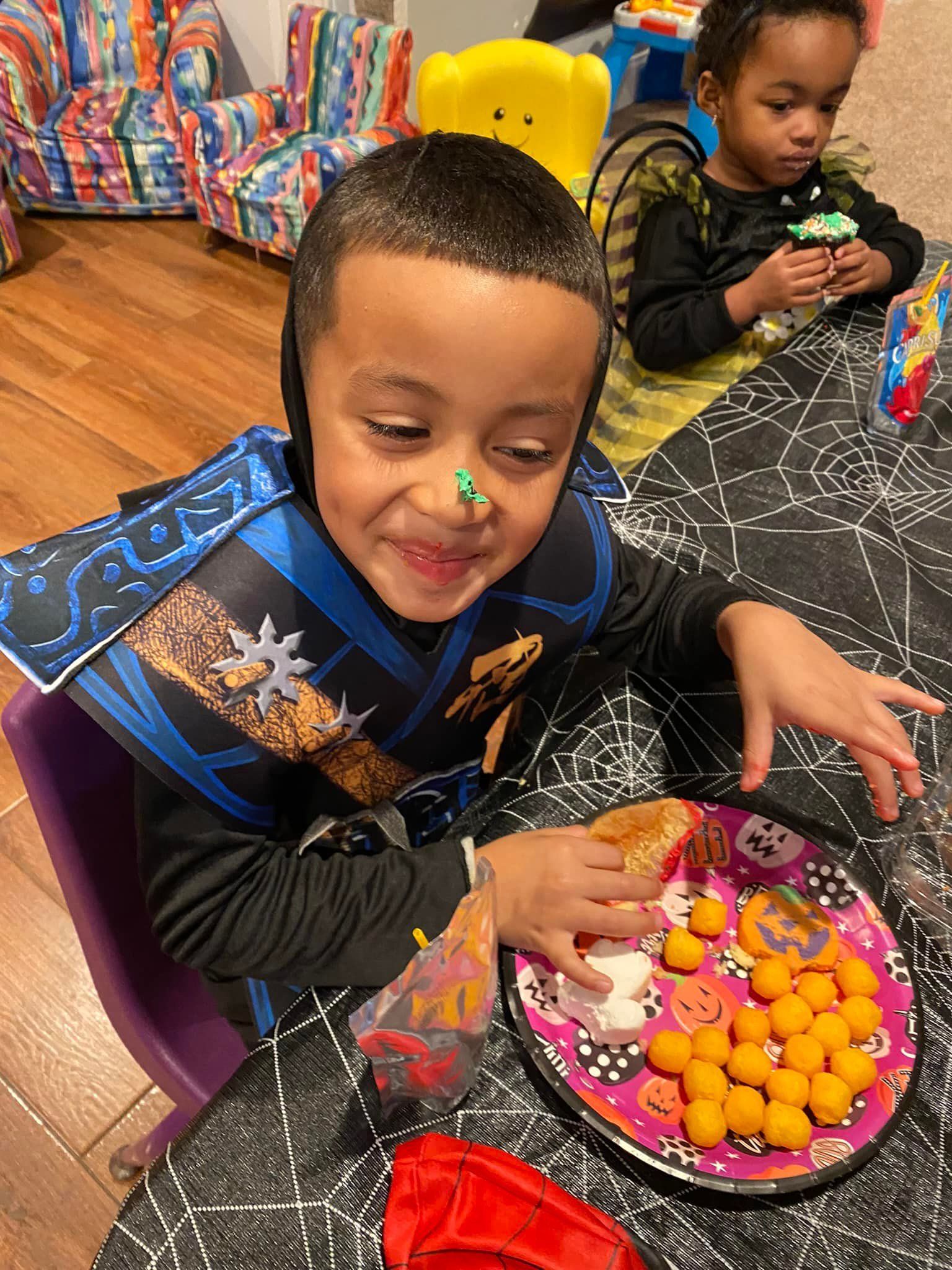 Child in a blue costume reaches for treats from a decorated bowl at a festive table, with another child in the background.