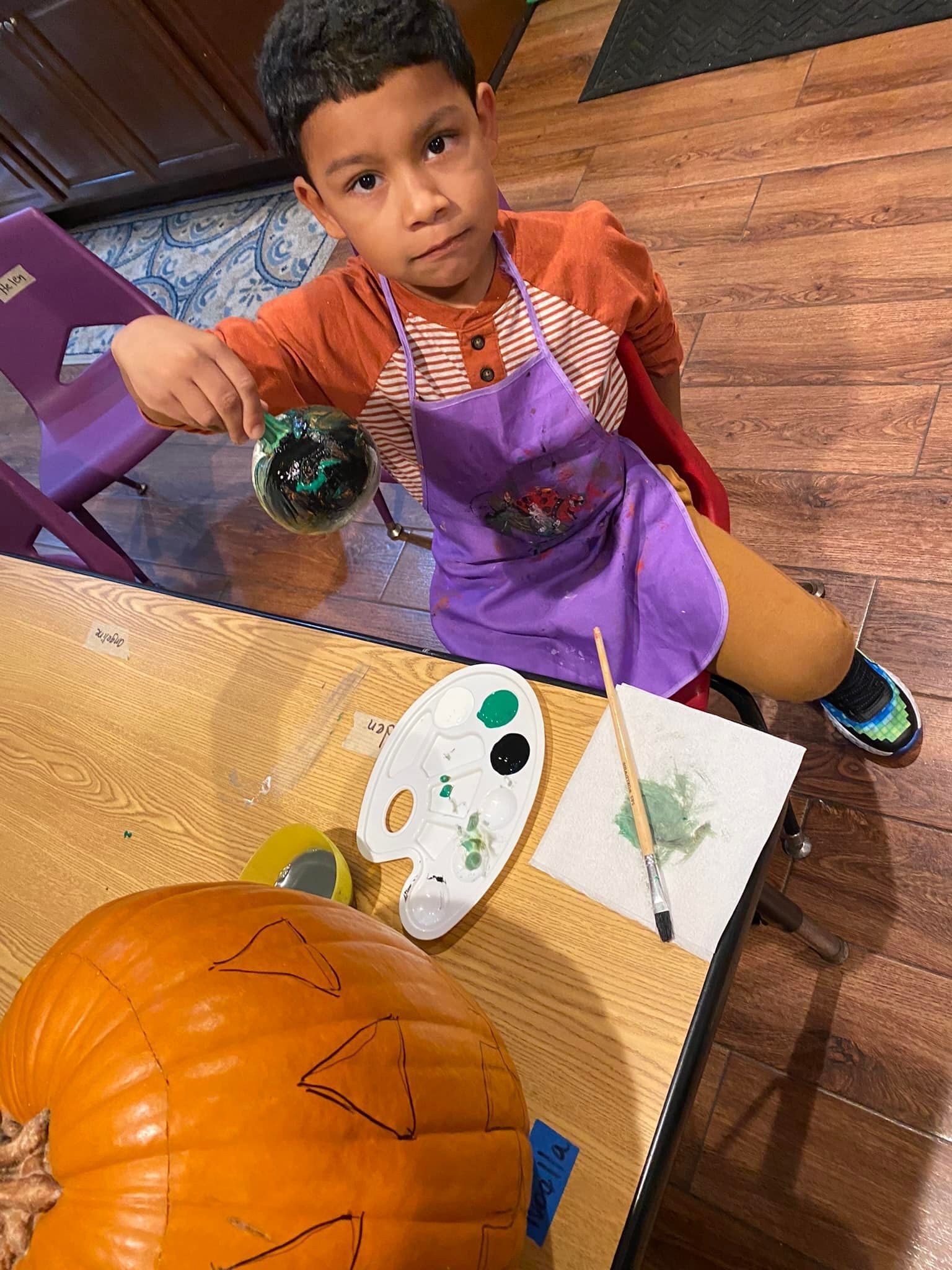 Child painting a pumpkin at a table with art supplies and a purple apron.