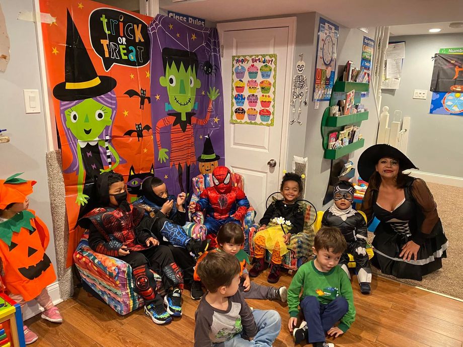 Costumed children in a Halloween-decorated classroom, with pumpkins, ghosts, and a witch decoration