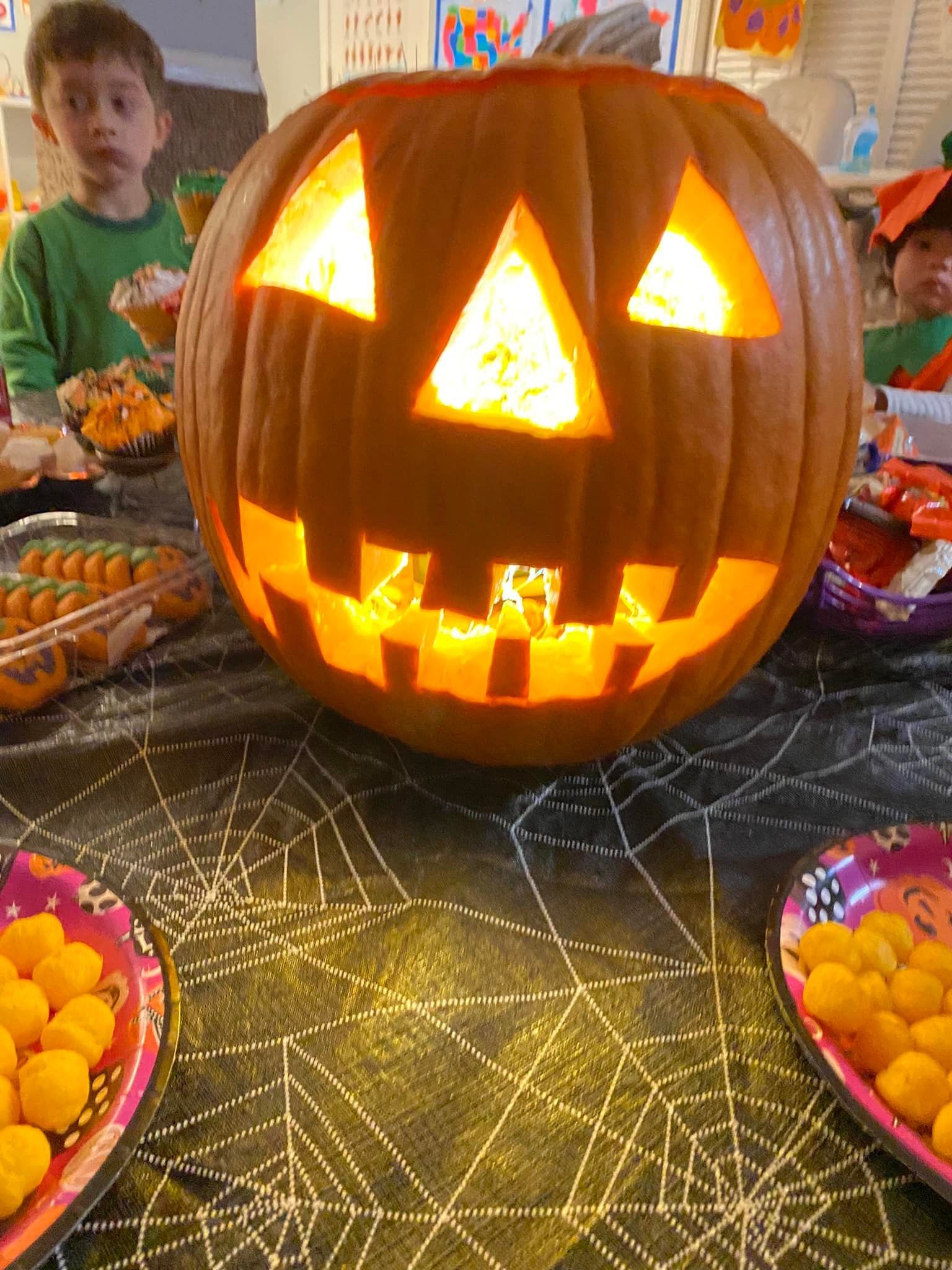 Lit jack-o’-lantern centerpiece on a Halloween table with candy bowls and children in the background