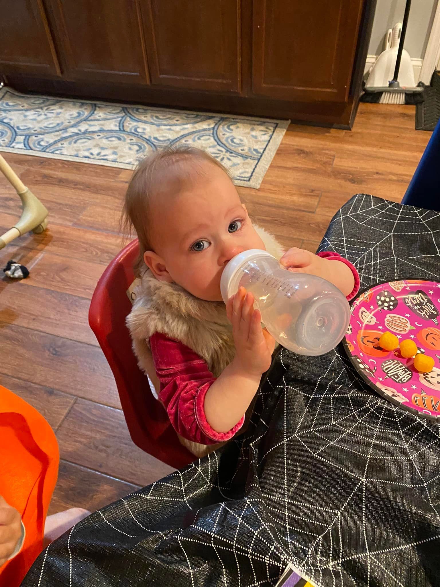 Child in a red chair drinking from a bottle at a table with a pink plate in a kitchen area
