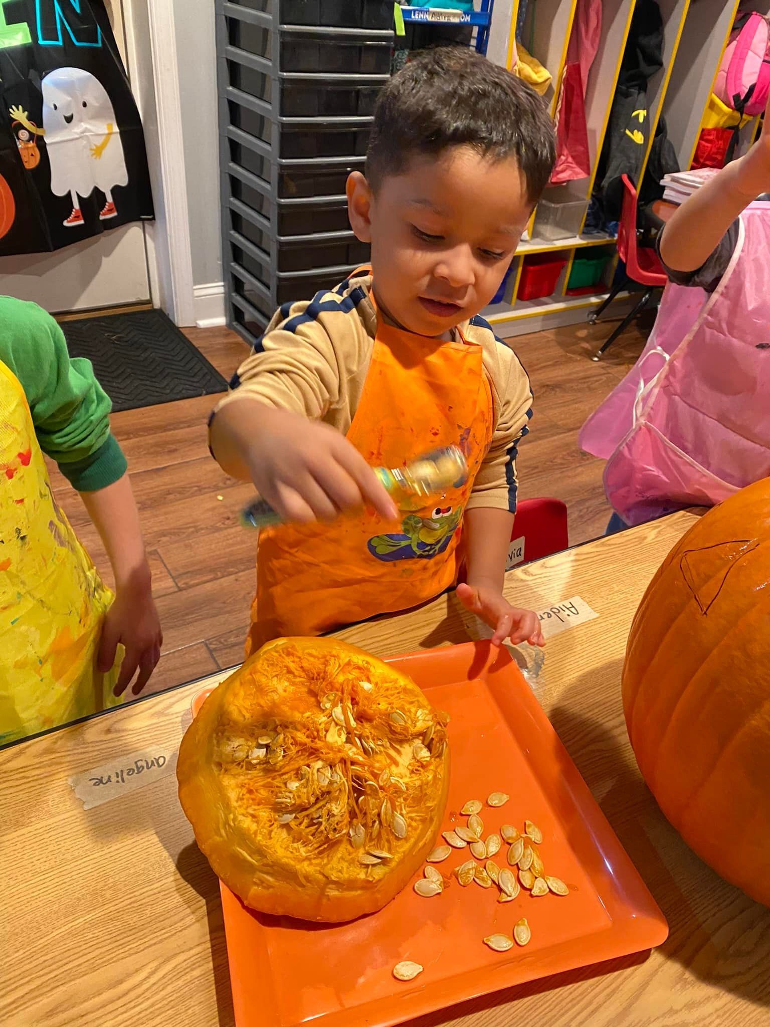 Child in orange shirt reaching for food on orange tray at a table, with a pumpkin nearby.