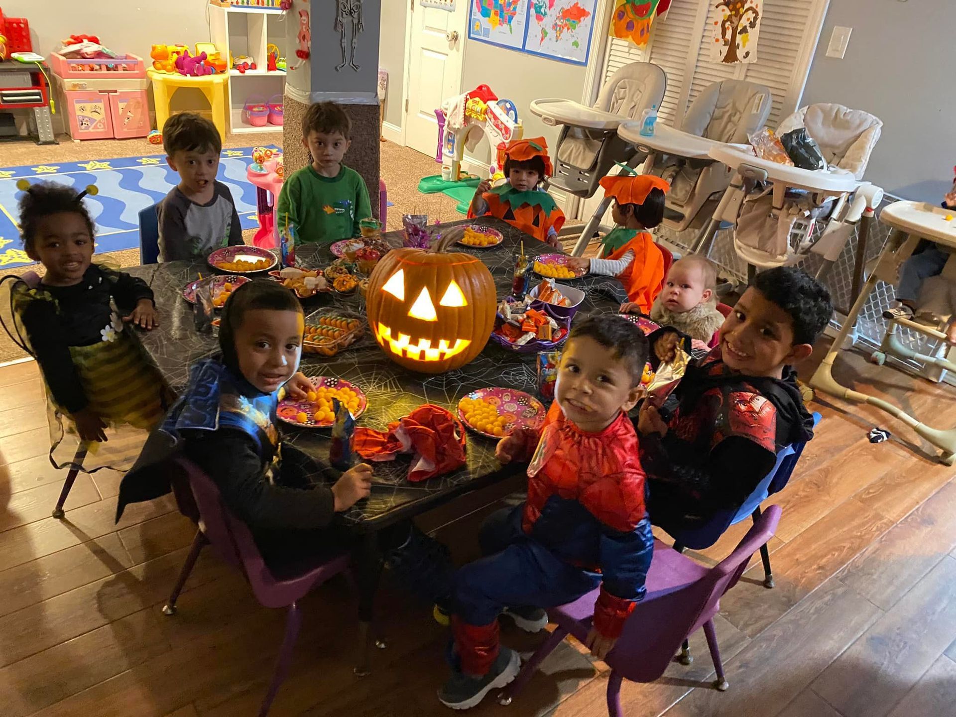 Children sit around a jack-o'-lantern centerpiece at a colorful classroom Halloween party.