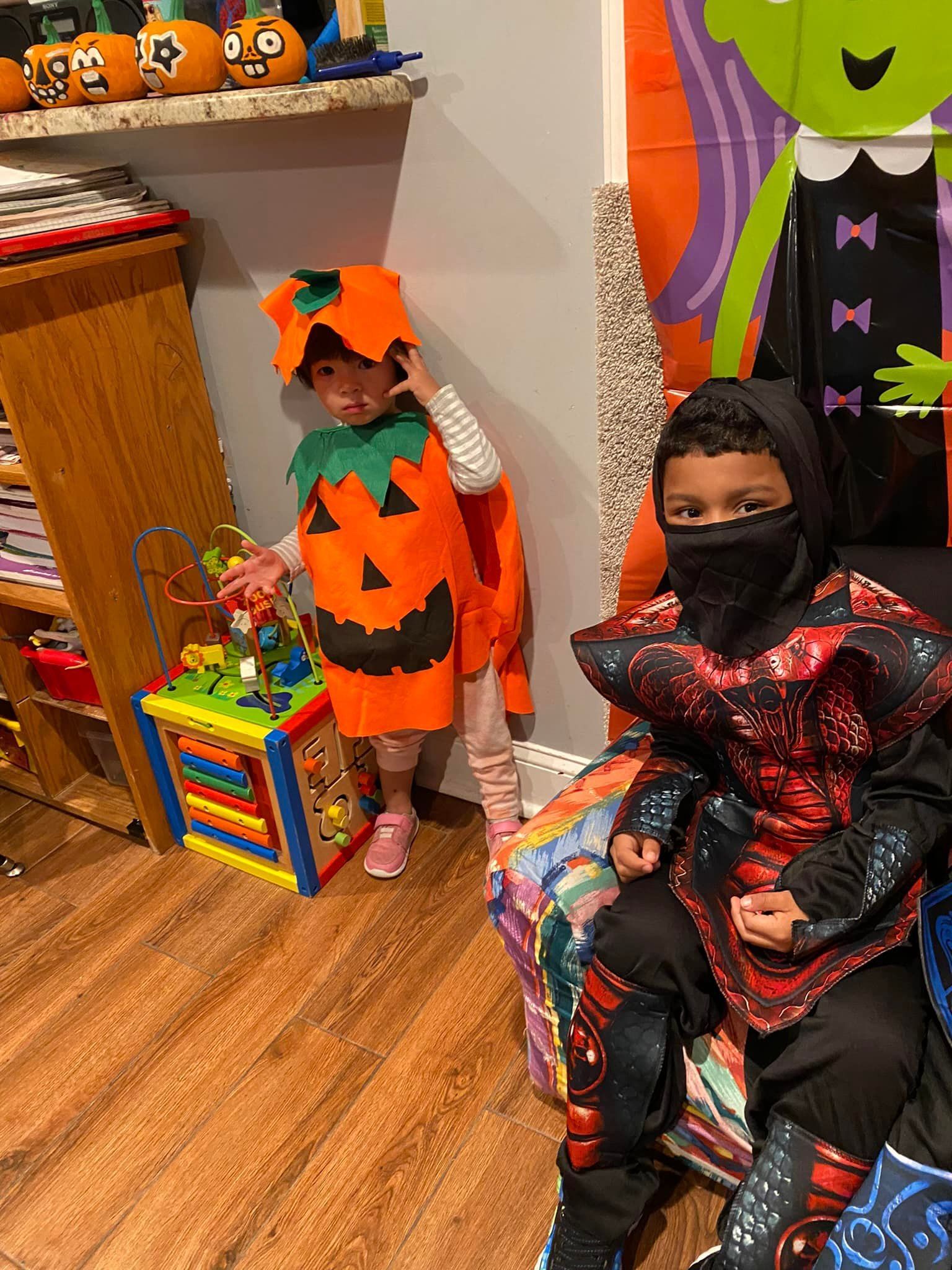 Two children in Halloween costumes pose indoors beside a pumpkin display and colorful decor.
