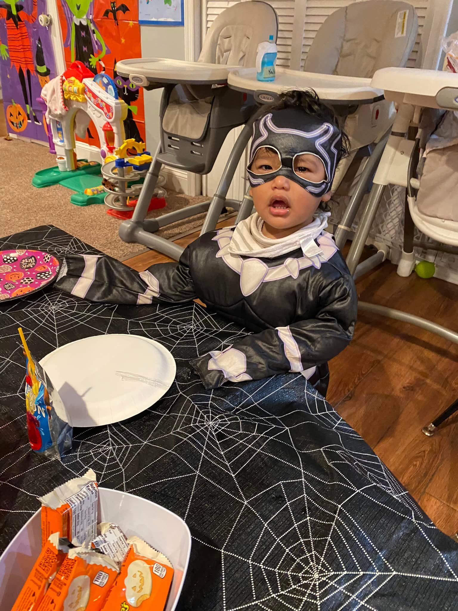 Child in a shiny black costume and mask sits on the floor at a party with a white plate nearby.