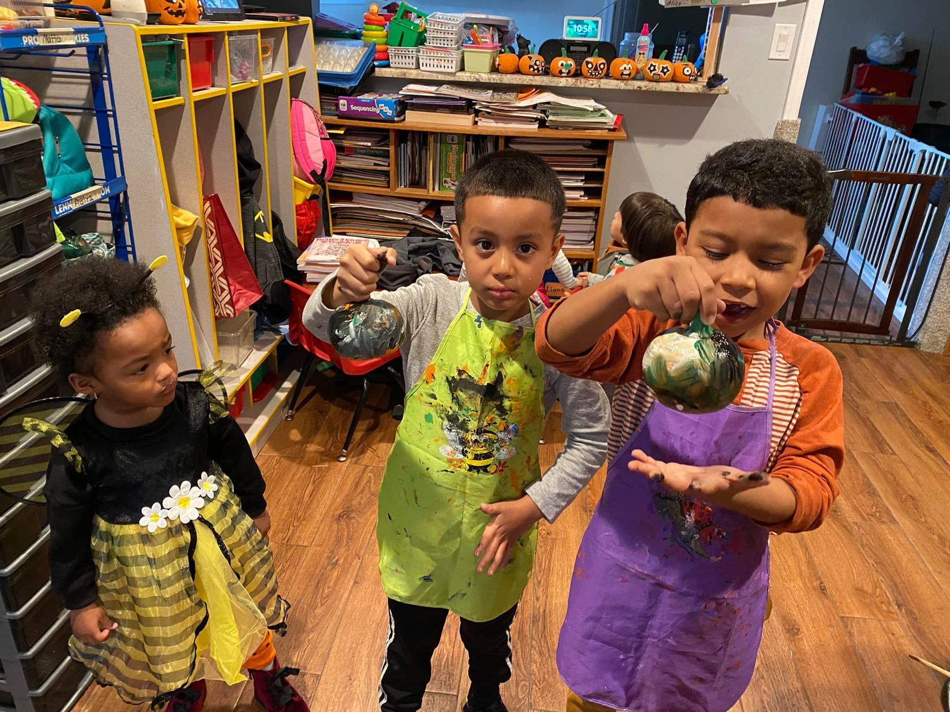 Three children in colorful aprons hold up painted pumpkins in a classroom.
