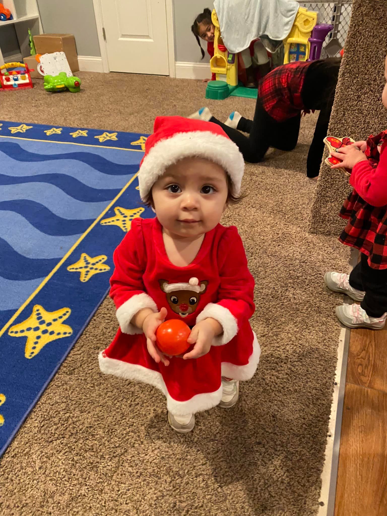 Toddler in a Santa outfit holding oranges in a classroom, standing on carpet near a blue rug.