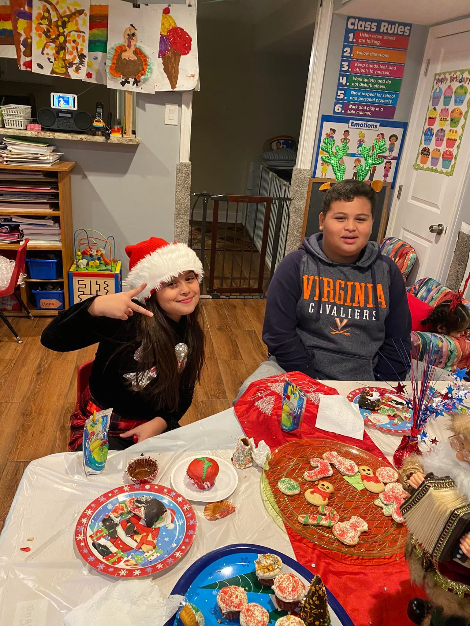 Two children sit at a Christmas craft table, smiling amid festive decorations and supplies.