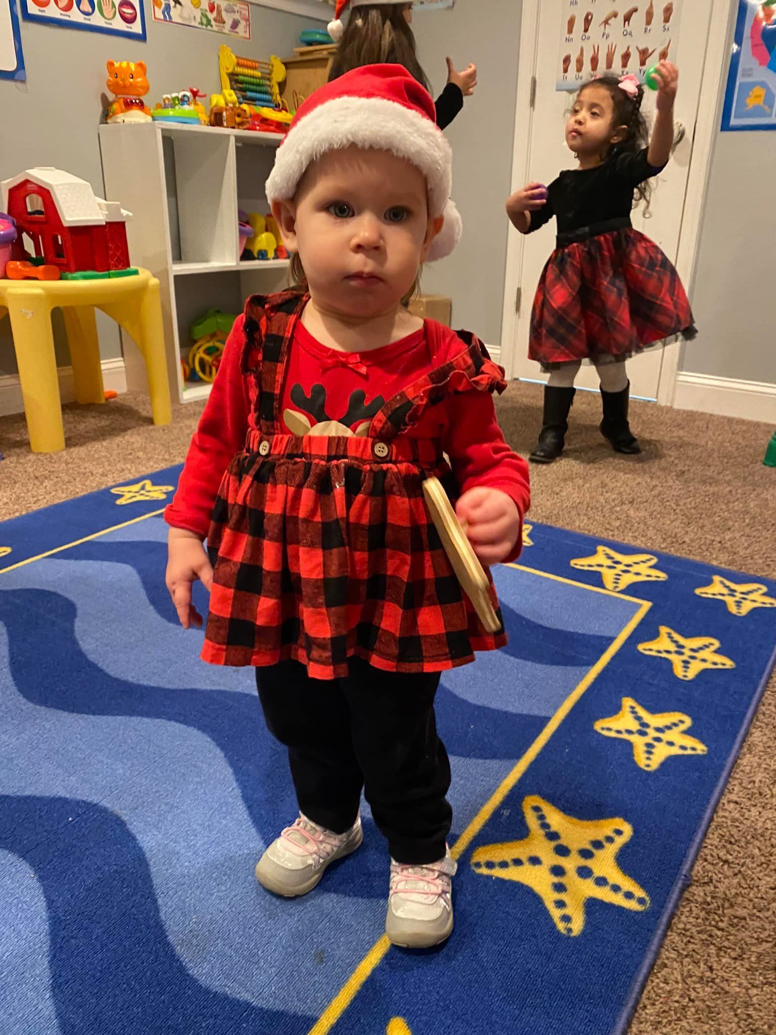 Toddler in red plaid dress and Santa hat standing on a blue star rug in a classroom, with another child in the background