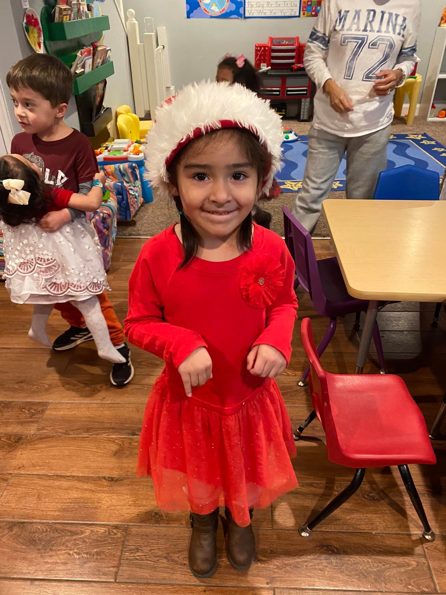 Child in a red dress and white hat smiles in a classroom, with toys and chairs around.