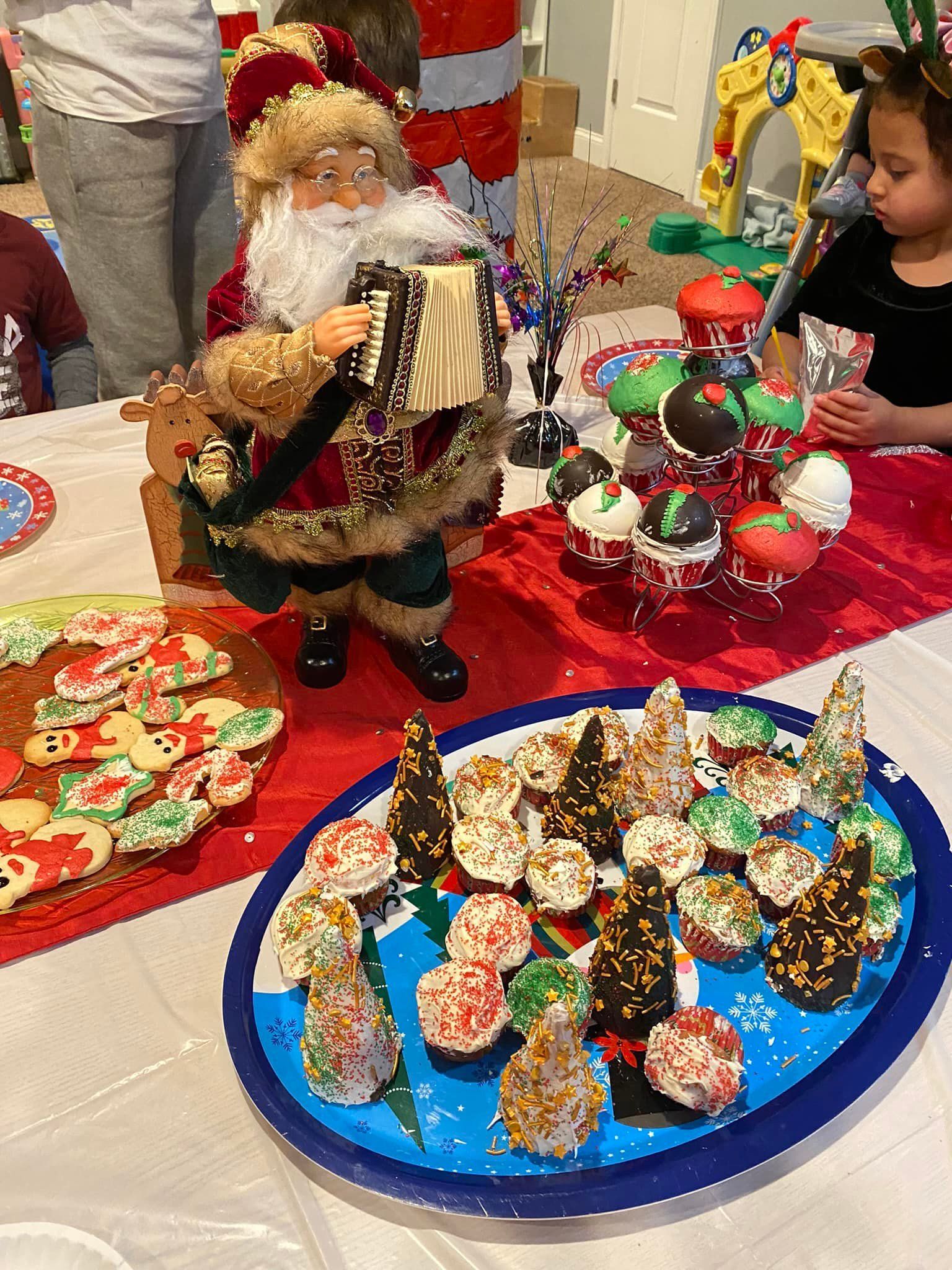 Santa figurine beside colorful Christmas cookie trays on a table, with a child reaching toward ornaments