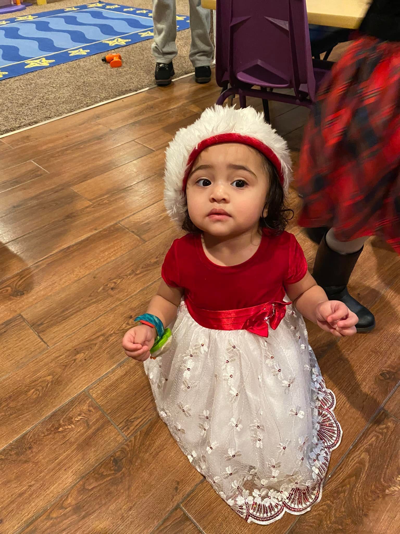 Child in a red-and-white dress and bonnet standing on a wood floor, looking up in a festive indoor setting