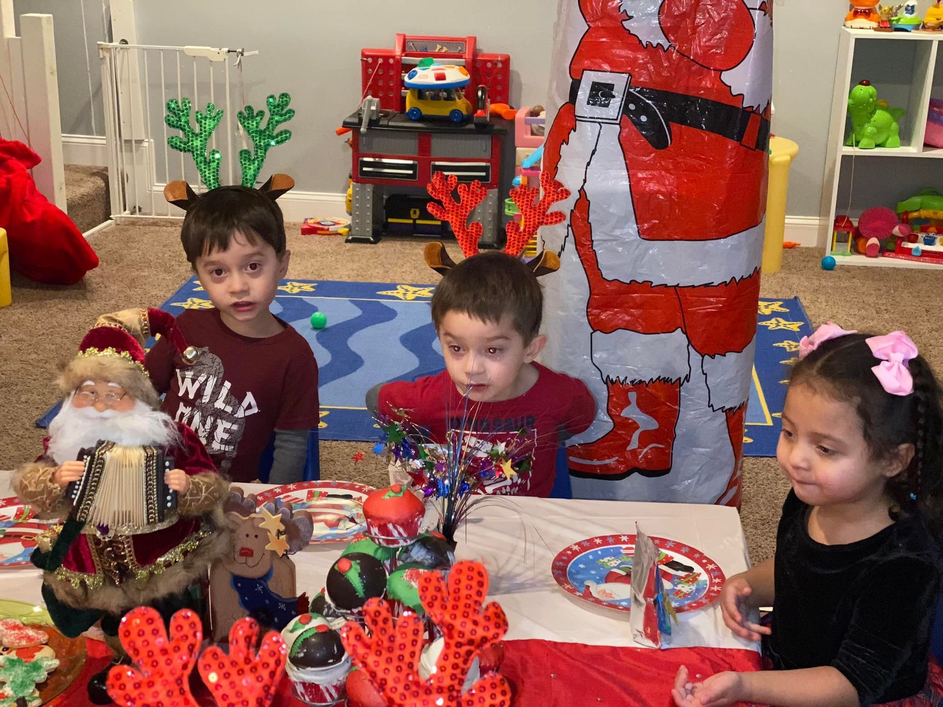 Children at a festive table with holiday decorations, crafts, and a Santa figure in the background