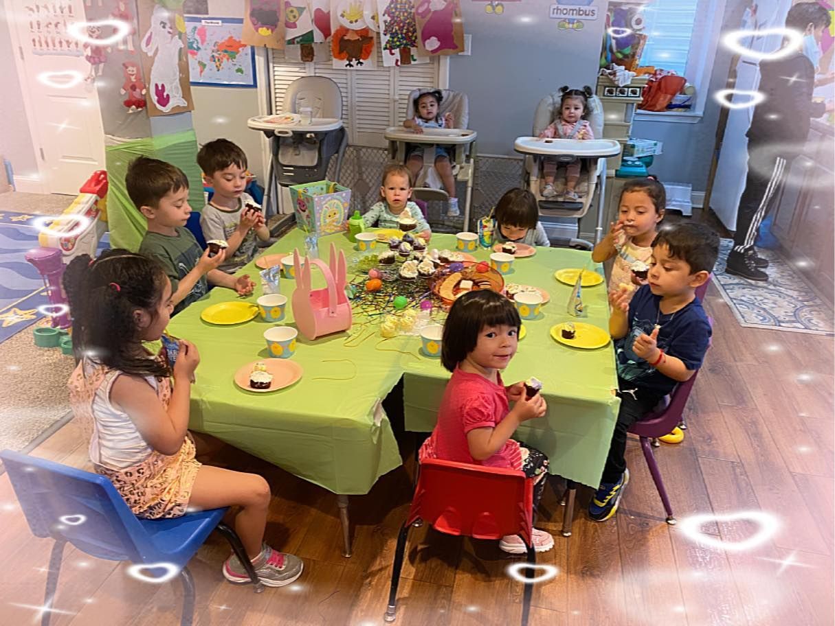 Children seated around a green table, eating snack in a bright classroom decorated with paper flowers.